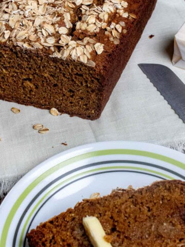 A buttered slice of Guinness brown bread on a plate with the loaf behind it.