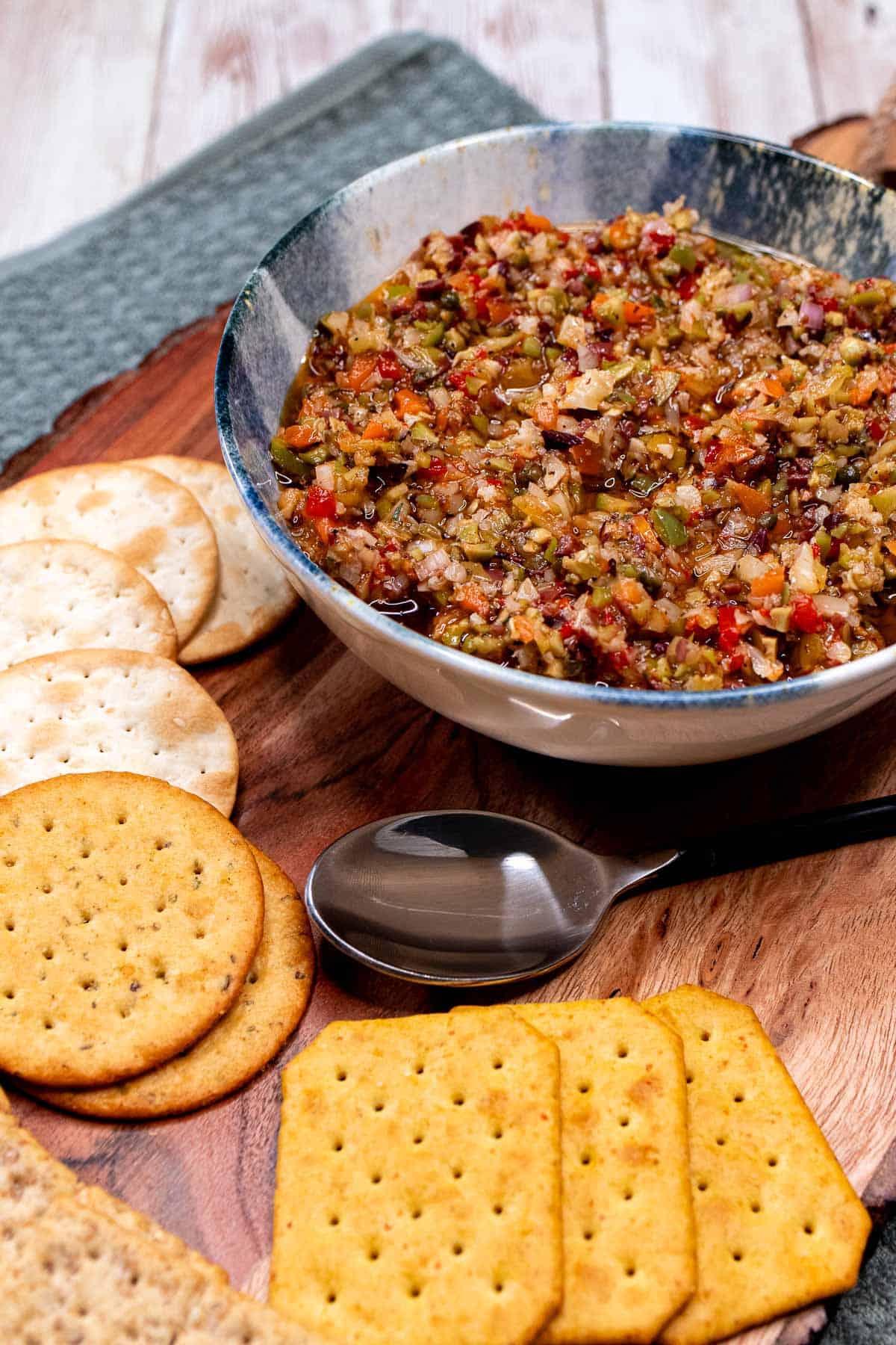 A bowl of olive salad served on a cheese board with assorted crackers.