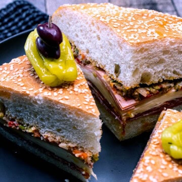 Sliced wedges of the New Orleans muffaletta sandwich on a serving plate, topped with a pepperoncini and Kalamata olives.