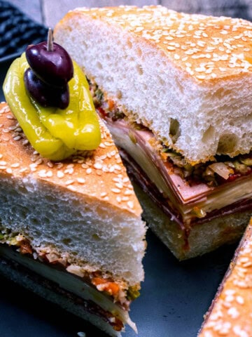 Sliced wedges of the New Orleans muffaletta sandwich on a serving plate, topped with a pepperoncini and Kalamata olives.