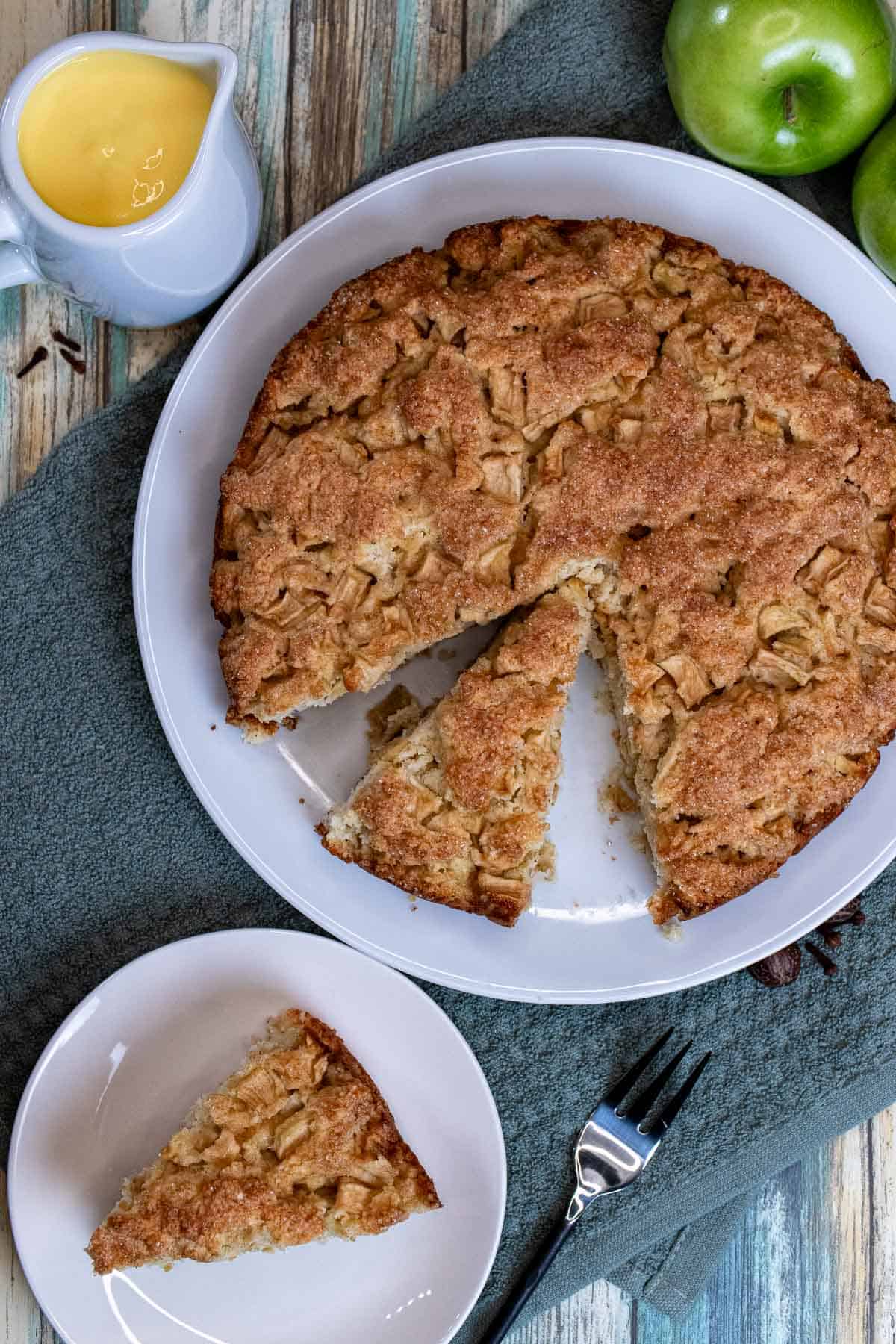 An Irish apple cake with vanilla custard sauce in a creamer pitcher and a cake slice on a small plate.