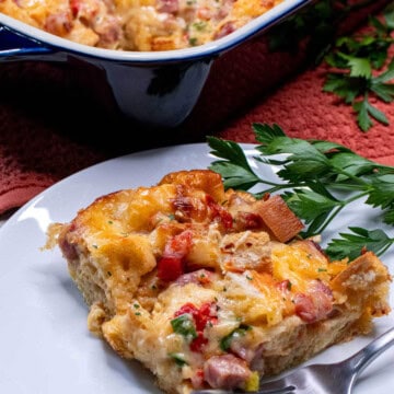 A slice of overnight ham and cheese strata on a plate with the baking dish behind it.