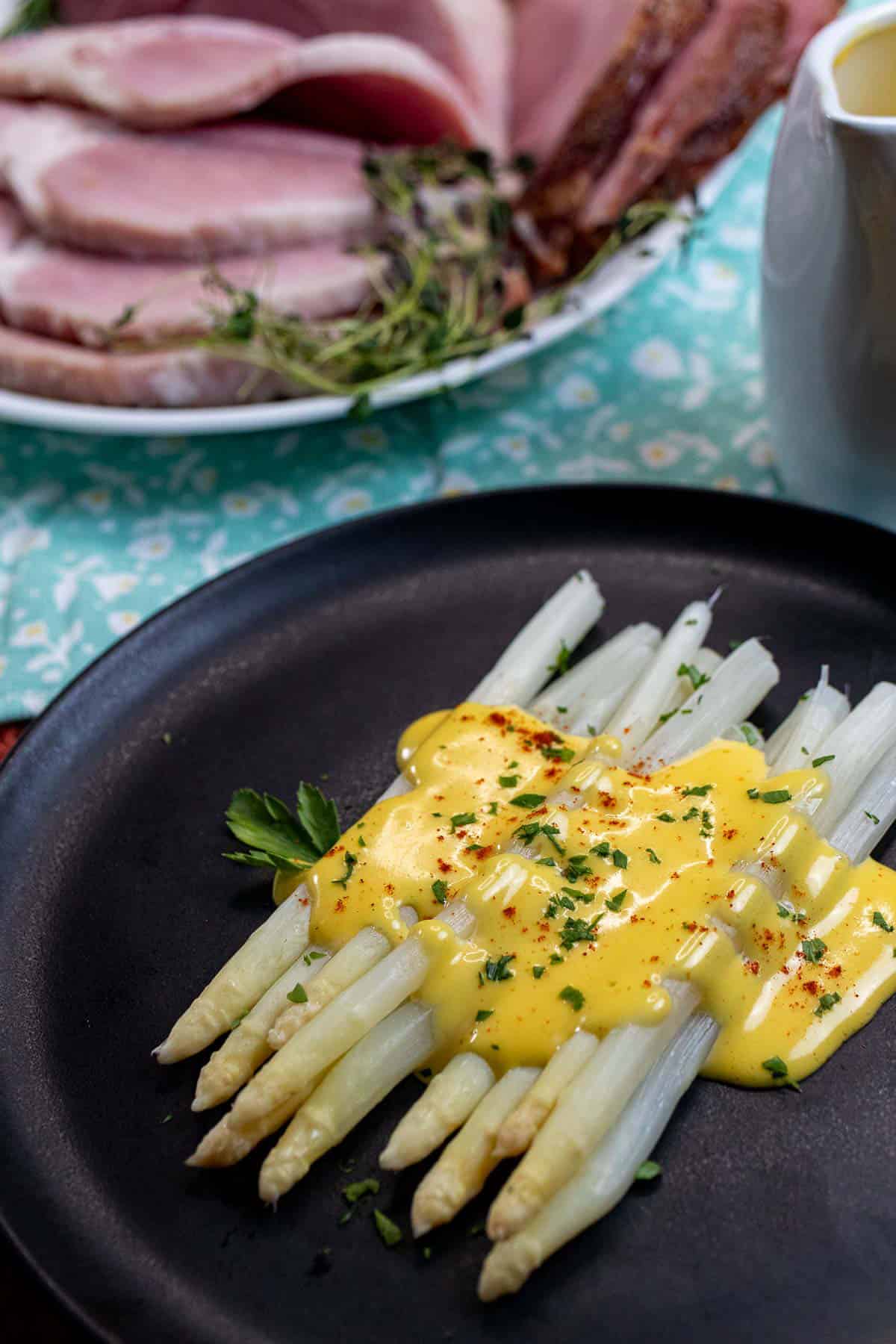 A plate of German white asparagus topped with hollandaise sauce with carved ham behind it.