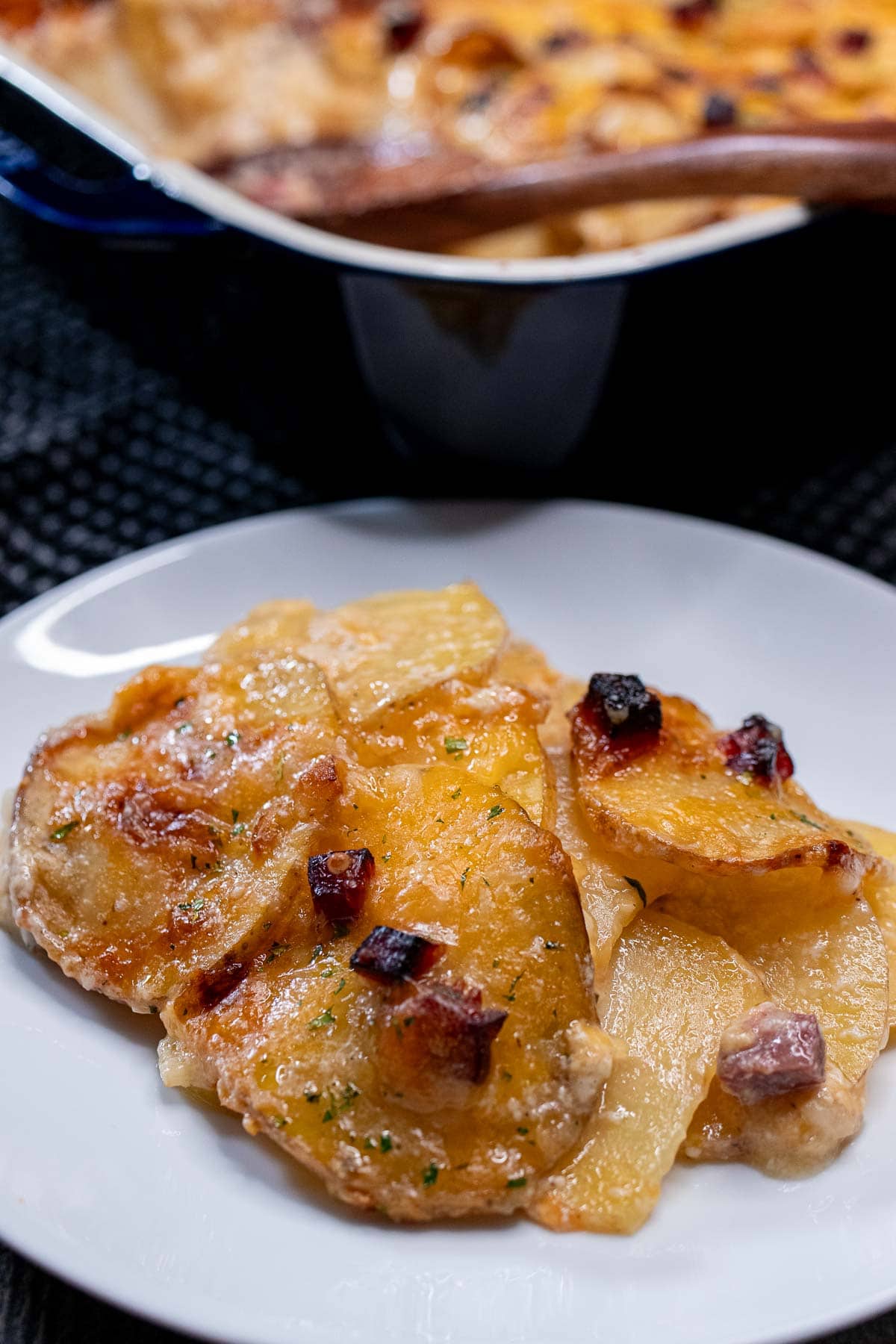 A small plate with a portion of cheesy scalloped potatoes with ham and the baking dish behind it.