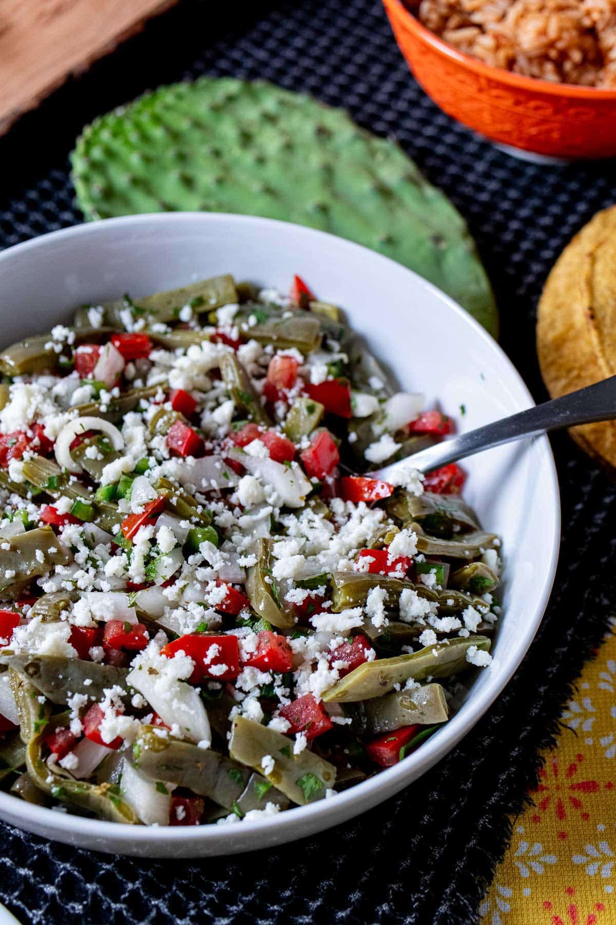 A serving bowl filled with ensalada de nopales, topped with crumbled queso fresco with a cactus pad behind it.