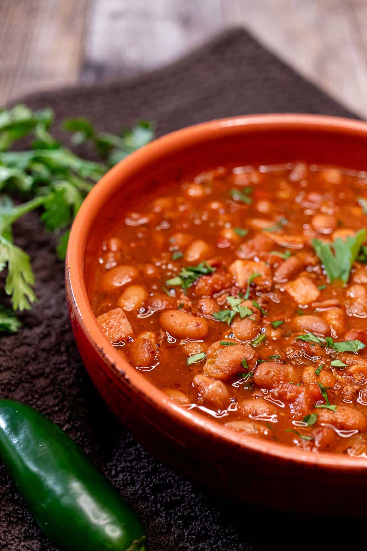 A bowl of frijoles charros topped with fresh cilantro with more cilantro and a jalapeno beside the bowl.