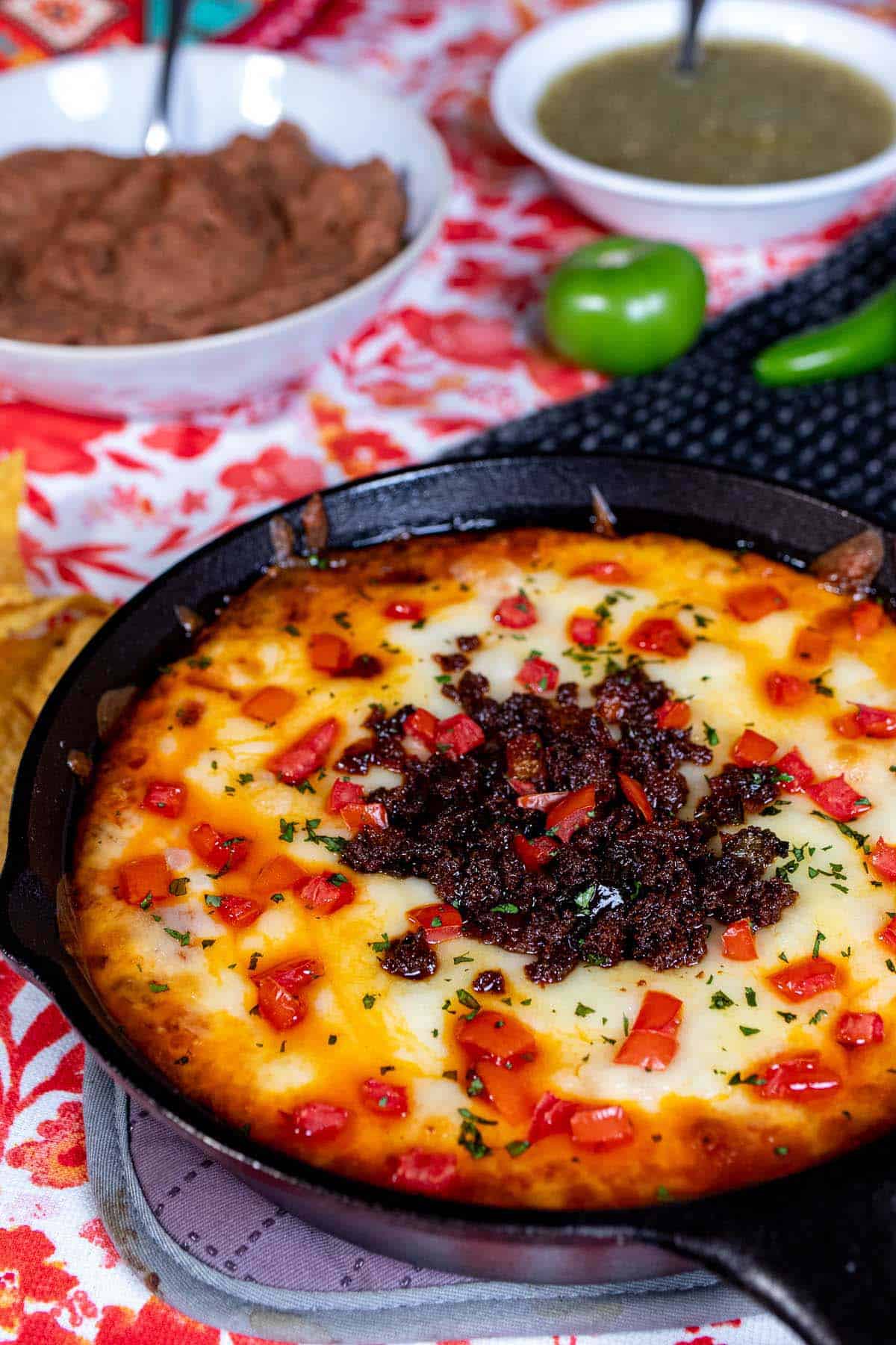 A skillet of queso fundido topped with chorizo, tomato, and cilantro with refried beans and salsa verde in the background.