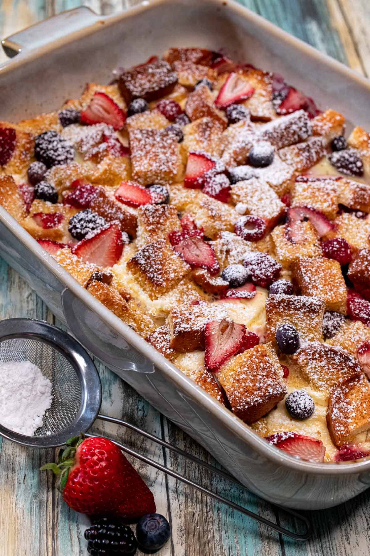 A full baking dish of overnight French toast casserole with mixed berries, with more fresh berries beside it.