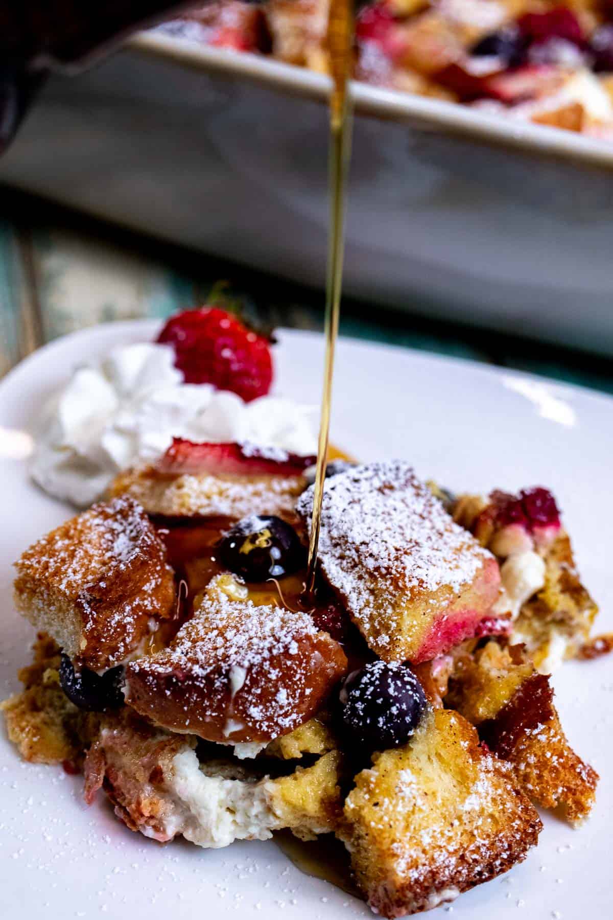 A portion of berries and cream French toast casserole with a side of whipped cream and being drizzled with maple syrup.