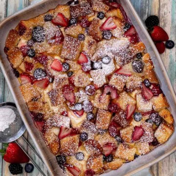A baking dish full of mixed berries and cream French toast casserole topped with powdered sugar with more fresh berries beside it.