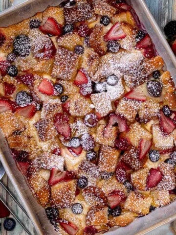 A baking dish full of mixed berries and cream French toast casserole topped with powdered sugar with more fresh berries beside it.