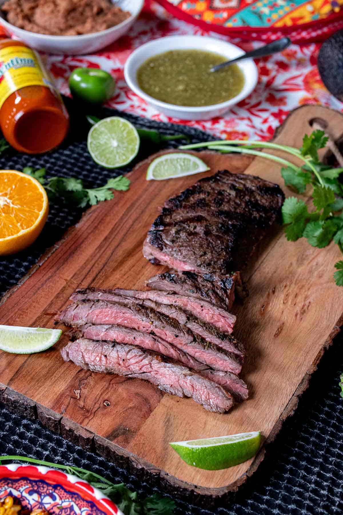 Sliced carne asada surrounded by cut orange and limes, cilantro, and salsas.