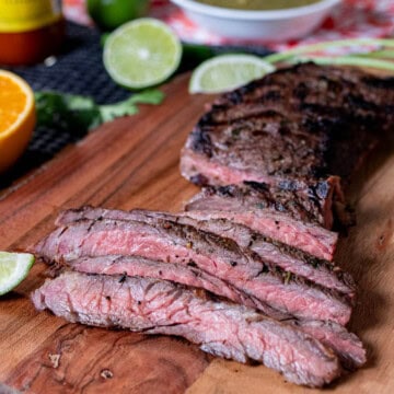 A sliced carne asada skirt steak on a wooden board, with cut limes and orange around it.