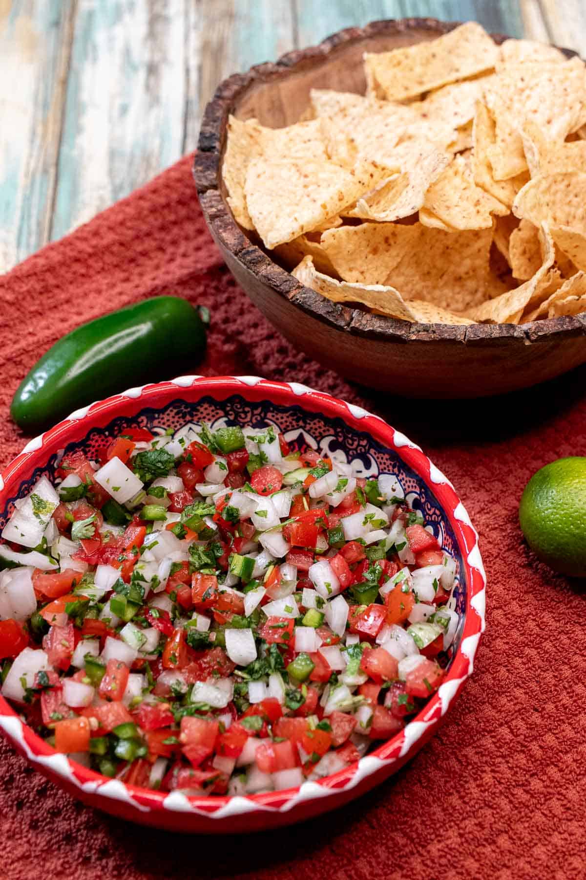 A red bowl full of fresh pico de gallo with a wooden bowl filled with tortilla chips behind it.