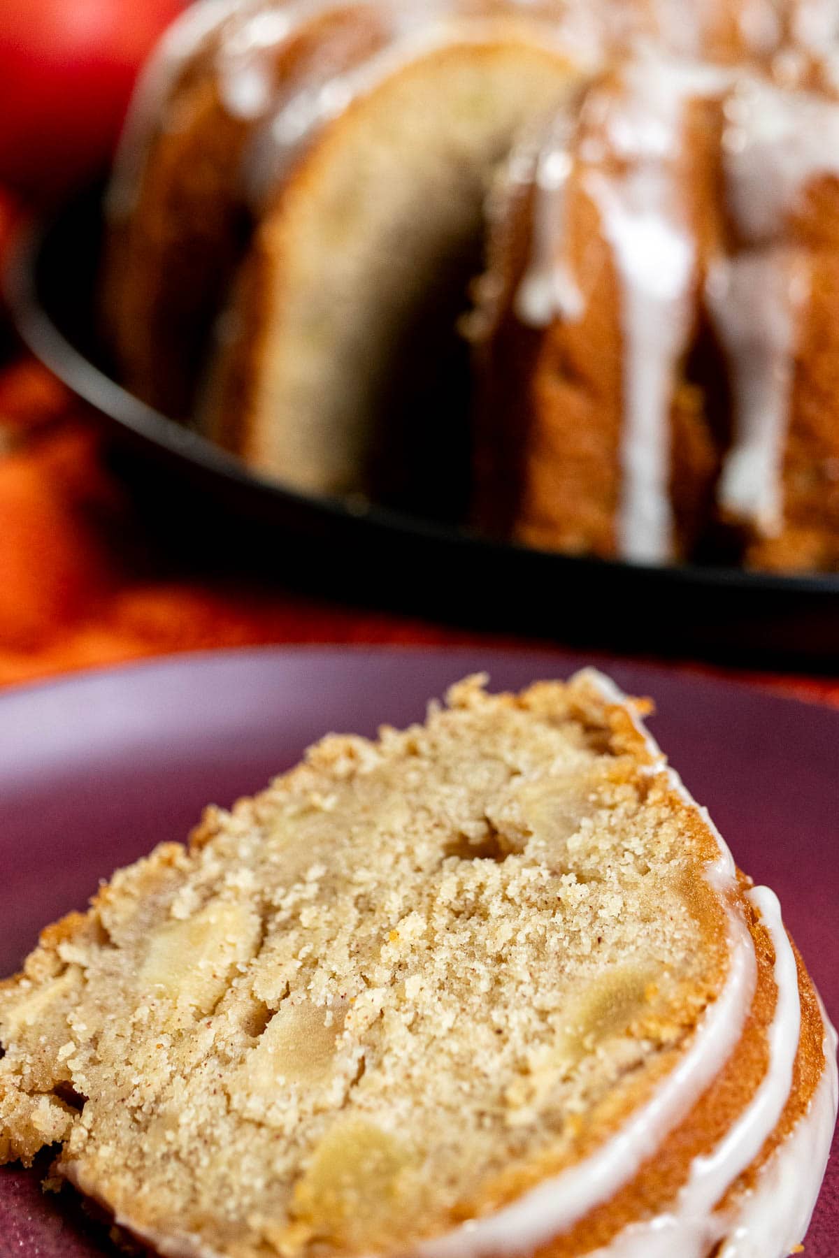 A slice of cider-glazed apple Bundt cake on a plate, with the rest of the cake behind it.