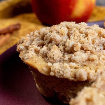 A cinnamon apple cider muffin with an apple and cinnamon sticks behind it.