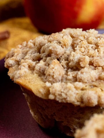 A cinnamon apple cider muffin with an apple and cinnamon sticks behind it.