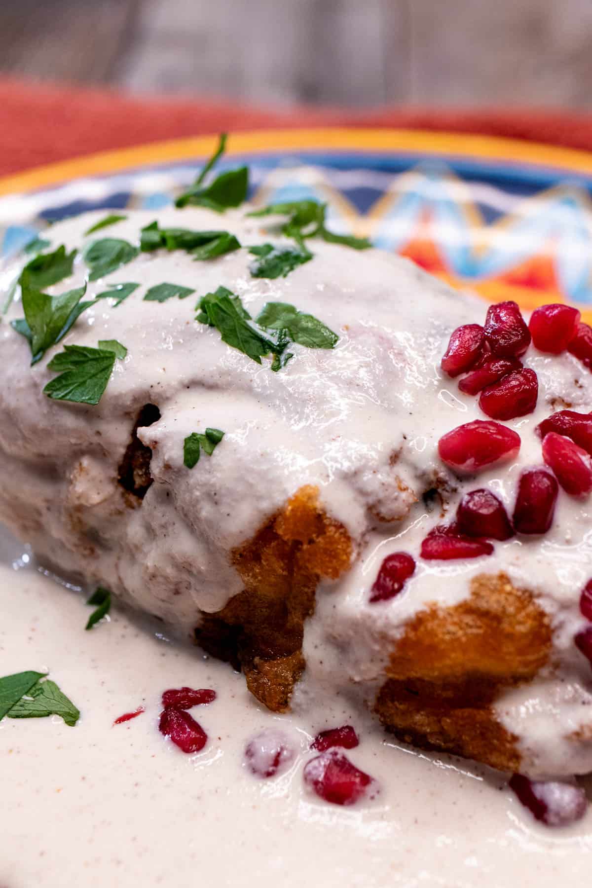 A chiles en nogada stuffed and fried on a plate covered with sauce and topped with parsley and pomegranate arils.
